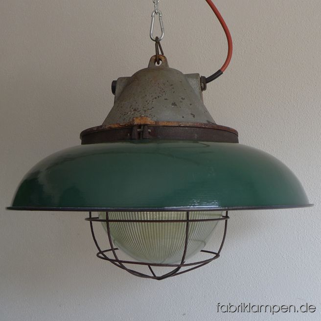 Sehr massive und schwere große Bunkerlampe mit Emailleschirm in grün. Diese Lampe ist DER Hammer: der Schirm hat einen Durchmesser von 52 cm, mit massiven gusseisernem Oberteil und Glaskolben wiegt die Lampe stolze 15 kg. Sie ist die größte ihrer Art und extrem selten zu finden. Diese Lampe besteht aus einem schweren gusseisernen Oberteil mit Schutzglas (geriffeltes Thermoglas) und Schutzgitter und einem grün (innen weiß) emaillierten Schutzschirm aus Stahlblech. Die alten Industrielampen haben die üblichen Spuren der vergangenen Jahrzehnte (Abplatzer, Kratzer, Verfärbungen, kleinere Roststellen, und Co.). Wir haben diese alle behandelt. Die Fabrikleuchten sind gereinigt und neu elektrifiziert (mit E27 Fassung). Abmessungen: Durchmesser 52 cm, Höhe 39 cm, Gewicht 15 Kg. Die Lampen werden mit 2 Meter Kabel geliefert (andere Längen sind natürlich möglich). Um diese massiven Industrielampen verwenden zu können, brauchen Sie eine tragfähige Decke – oder starke Balken, die diese recht gewichtigen Leuchten  halten können. 