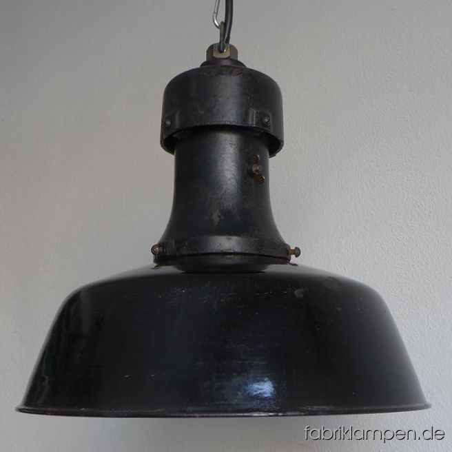 Antike schwarze Fabriklampe mit Emailleschirm und gusseisernem Oberteil, ca. aus den 1920-ern Jahren. Die Lampe hat die sehr starken Spuren der vergangenen Jahrzehnte (Abplatzer, Roststellen, Kratzer, Verfärbungen und Co. – wir haben die alle behandelt). Sie ist gereinigt und neu elektrifiziert, mit E27-Keramikfassung. Material: schwarz (innen weiß) emailliertes Stahlblech mit ausgeprägten Gebrauchsspuren, gusseisernes Oberteil. Sie bekommen diese Lampe mit 2 Meter Kabel und massiver Aufhängeöse für die sichere Aufhängung. Gegen Aufpreis kann die Lampe auch mit Ketten-Aufhängung geliefert werden. Die Abmessungen: Durchmesser Lampe (Schirm) 40 cm, Höhe 37 cm. 